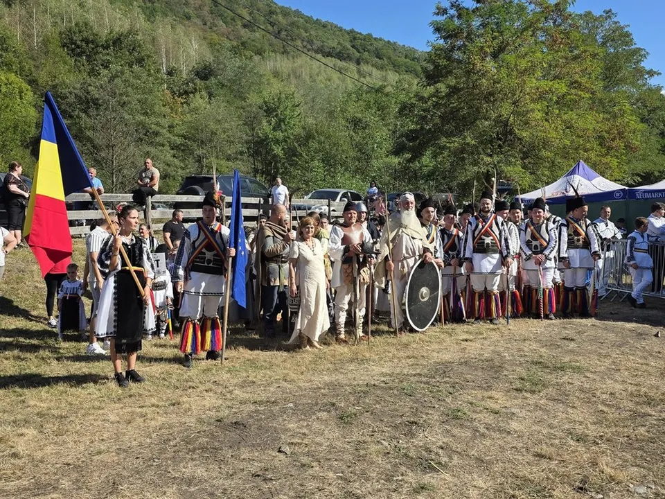 Un grup numeroas în costume tradiționale stă pe iarbă, lângă steaguri, scuturi și un cort, cu un deal împădurit în fundal.