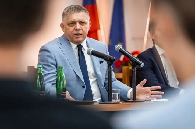 Un bărbat într-un costum albastru deschis și cravată închisă stă la o masă cu microfoane și sticle de apă în fața unor steaguri.