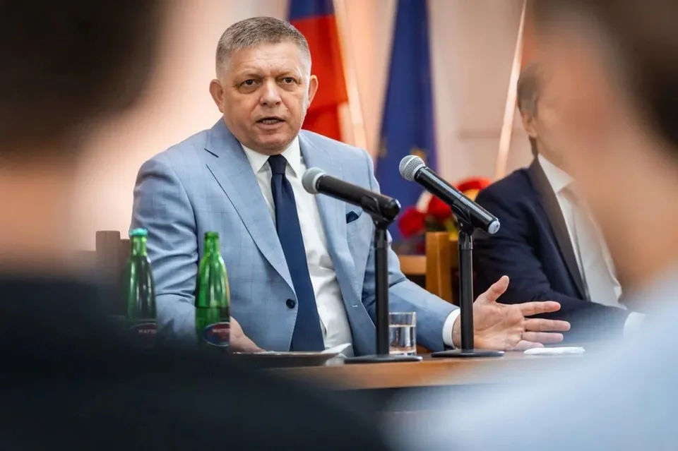 Un bărbat într-un costum albastru deschis și cravată închisă stă la o masă cu microfoane și sticle de apă în fața unor steaguri.