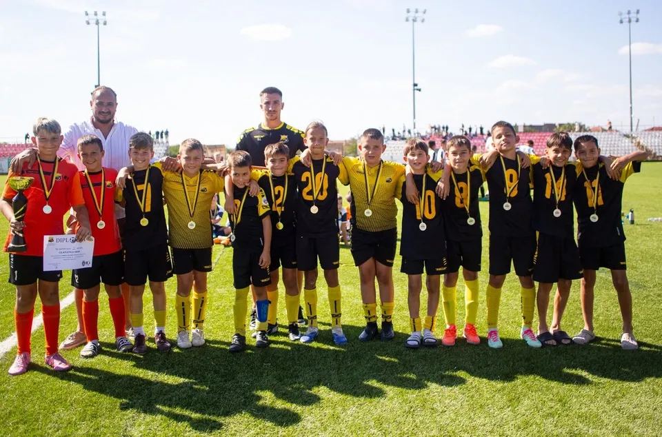 Un grup de copii îmbrăcați în echipamente sportive negre și galbene stau pe un teren de fotbal, purtând medalii aurii în jurul gâtului.