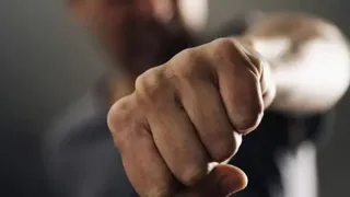 Un pumn închis este poziționat în prim-plan, în timp ce o persoană este vizibilă într-un fundal neclar și desfocat.