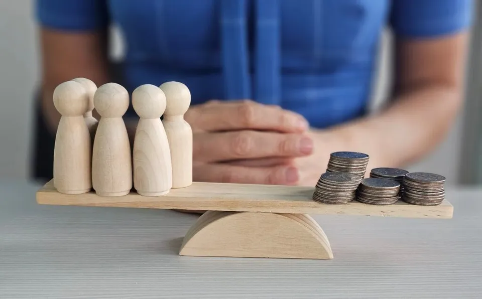 Cinci figurine de lemn stau pe o parte a unei balanțe de lemn, în timp ce stive de monede se află pe cealaltă parte.