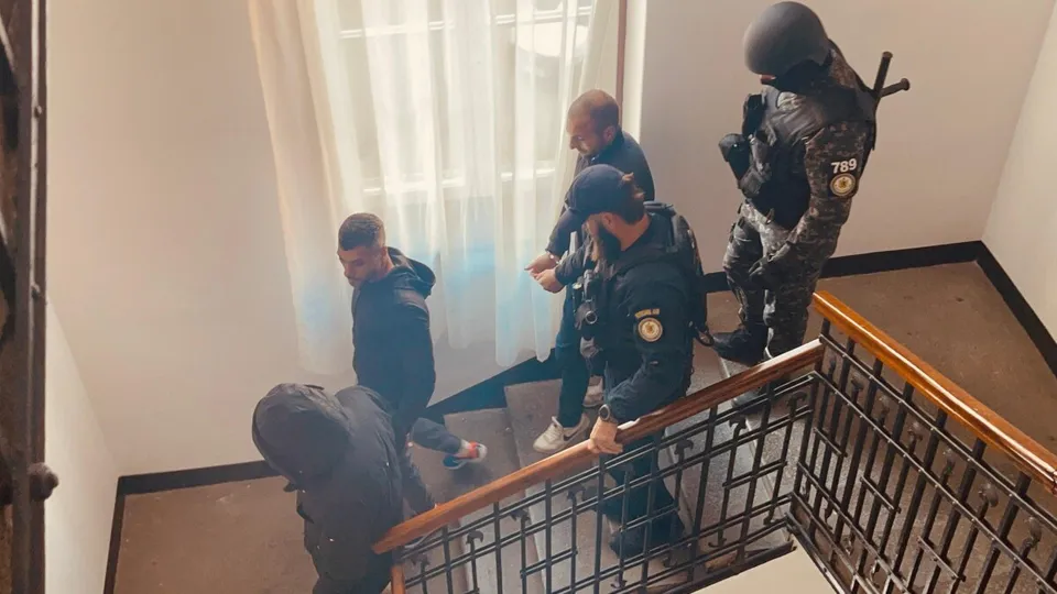 Patru bărbați, dintre care unul poartă uniformă de polițist și echipament tactic, coboară pe o scară interioară lângă un balustradă metalică.