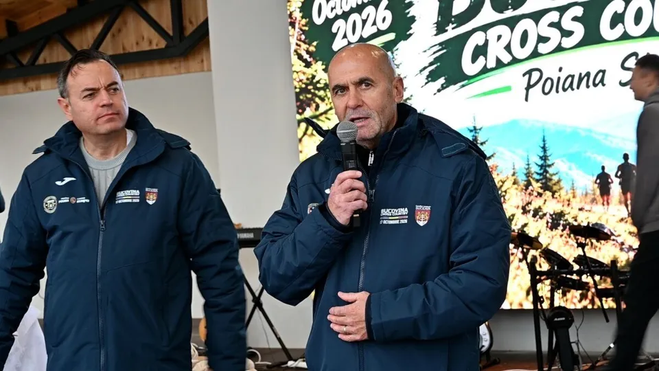 Un bărbat cu ochelari și o jachetă albastră ține un microfon în mână, în timp ce un alt bărbat în jachetă albastră stă lângă el.