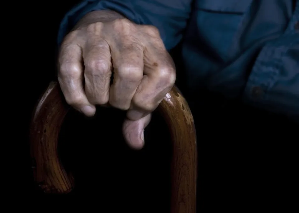 O mână bătrână cu pielea ridată este strânsă pe mânerul de lemn al unui baston pe un fundal negru.