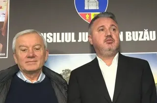 Un bărbat cu părul alb poartă un pulover albastru și o jachetă neagră, stând lângă un bărbat cu părul scurt, într-un sacou negru și cămașă albă.