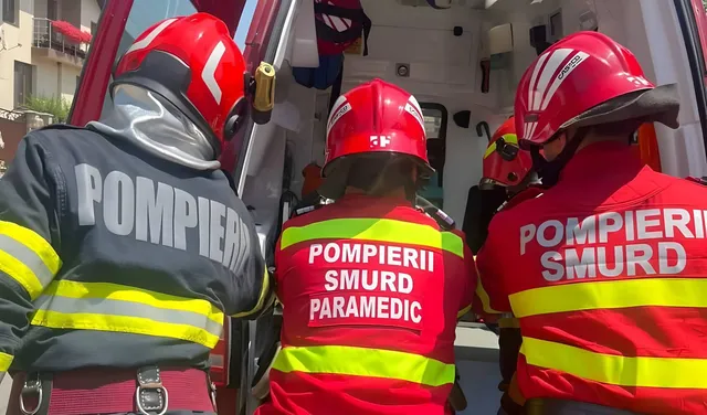 Trei persoane purtând uniforme de pompieri și căști de protecție roșii stau cu spatele în fața unui vehicul de intervenție.