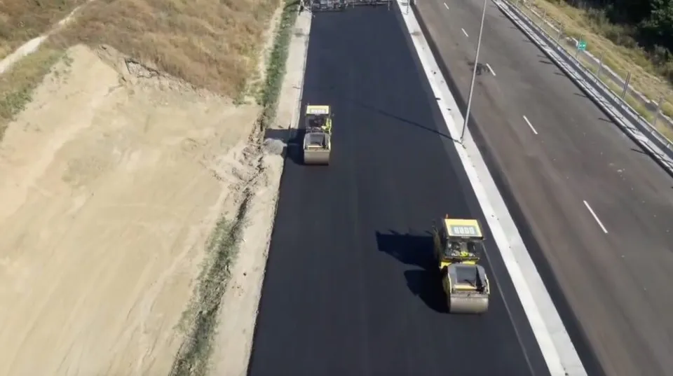 Două compactoare galbeni aspiră un drum nou, asfaltat, alăturate pe o bandă de circulație.