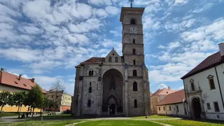 O biserică din piatră cu turn, poartă arcuită și ferestre înguste stă într-o curte cu iarbă, clădiri laterale, copaci și cer noros.