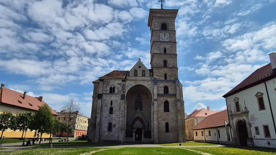 O biserică din piatră cu turn, poartă arcuită și ferestre înguste stă într-o curte cu iarbă, clădiri laterale, copaci și cer noros.