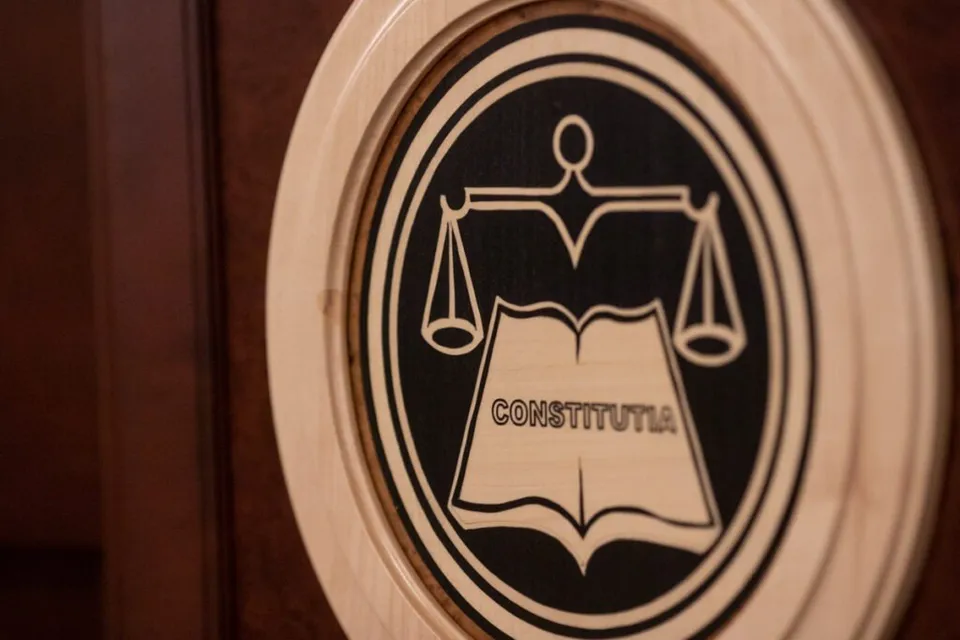Un emblema circulară din lemn încadrată într-un cerc, reprezentând o balanță deasupra unei cărți deschise pe care scrie textul CONSTITUTIA pe un fundal negru.