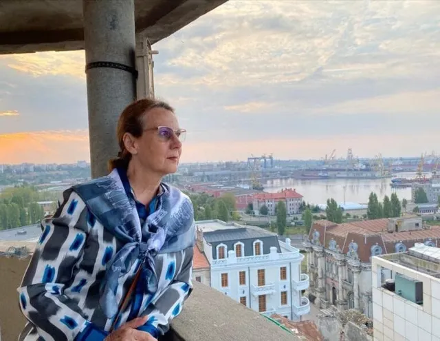 O femeie cu ochelari și o bluză modelată cu dungi albastre și albe stă pe un balcon cu orașul și un port în fundal.