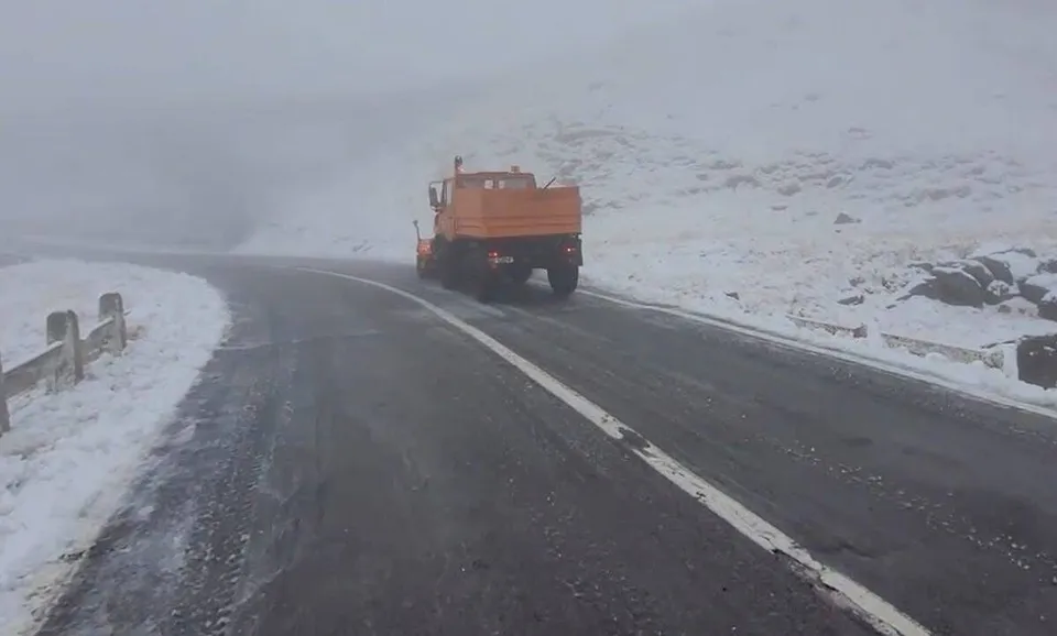 Un camion portocaliu circulă pe un drum asfaltat cu linii albe, înconjurat de un peisaj acoperit de zăpadă sub un cer înnorat.