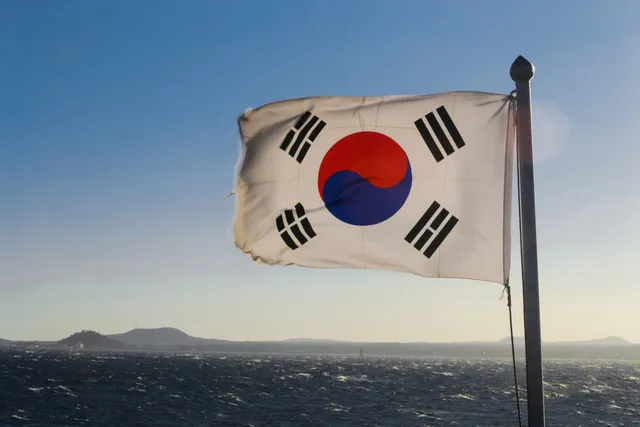 O steag sud-coreean flutură pe un stâlp sub un cer albastru, deasupra unei mări cu valuri mici și unor munți în depărtare.