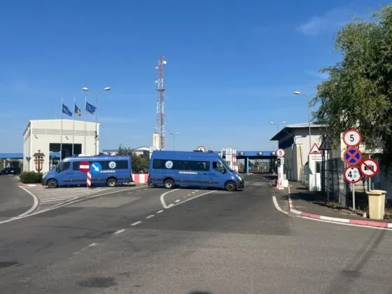 Două microbuze albastre sunt parcate pe un drum asfaltat lângă clădiri albe sub un cer senin.