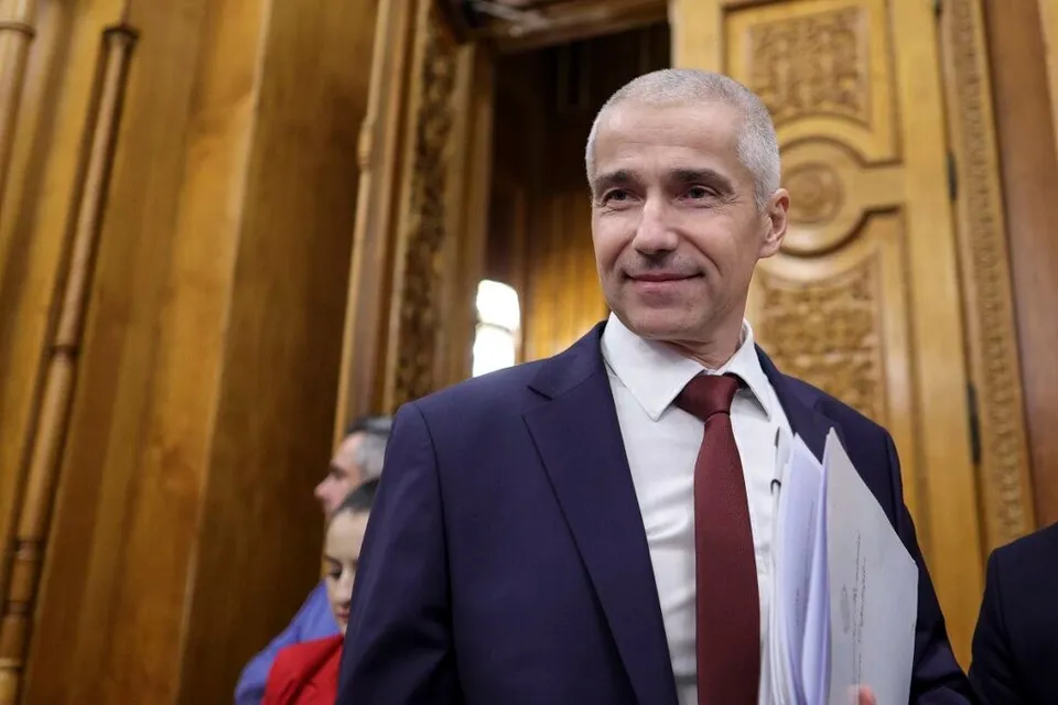 Un bărbat cu părul scurt și gri, purtând sacou închis la culoare, cămașă albă și cravată roșie, ține în mână un dosar alb.