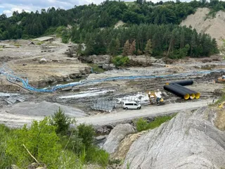 Un teren accidentat cu pământ, pietre, conducte negre și albastre, un vehicul alb și utilaje de construcții sub un cer înnorat.