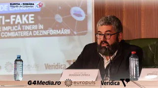 Un bărbat cu barbă și ochelari stă la o masă lângă două sticle de apă, în fața unui ecran de proiecție.