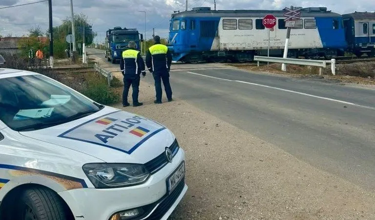 Două persoane în uniforme galben și albastru stau pe marginea unui drum lângă un tren albastru și un vehicul de poliție alb.