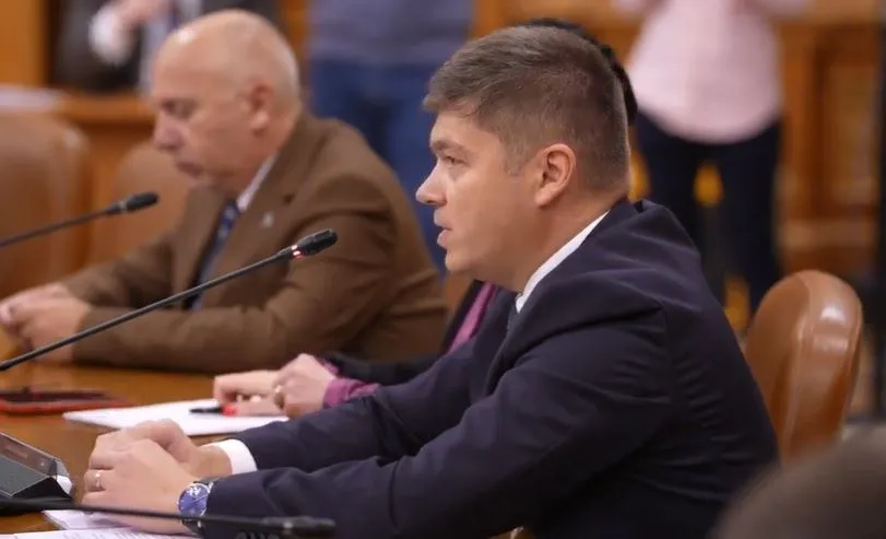 Un bărbat cu părul scurt și costum închis stă la o masă cu microfoane, în timp ce un alt bărbat cu costum maro este așezat în fundal.
