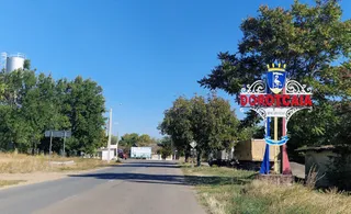 Un indicator stradal cu un blazon colorat stă pe marginea unei drumuri asfaltate lângă copaci sub un cer senin.