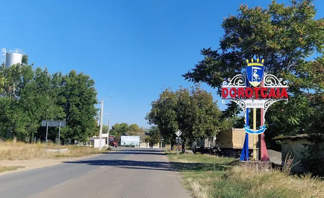 Un indicator stradal cu un blazon colorat stă pe marginea unei drumuri asfaltate lângă copaci sub un cer senin.