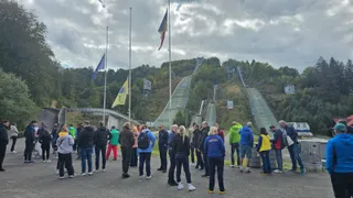 Un grup de persoane stă pe o suprafață asfaltată în fața unor săli de sărituri cu ski în mijlocul unei păduri sub un cer înnorat.