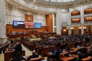 O sală de ședințe mare cu mulți oameni așezați la birouri de lemn sub un plafon înalt și decoruri de perete cu panouri de lemn.