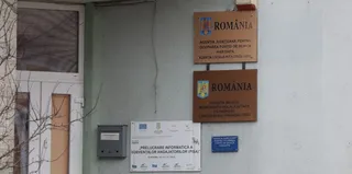 Două plăcuțe maro cu inscripții sunt montate pe un perete alb lângă o ușă de culoare albă.