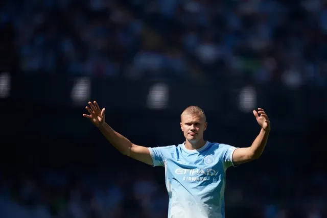 Un bărbat cu părul scurt și deschis la culoare poartă un tricou albastru deschis și stă cu ambele brațe întinse lateral, palmele orientate spre înainte.