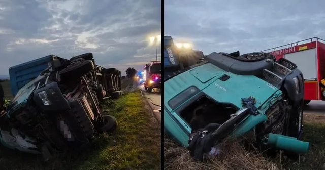 Un camion cu remorcă este răsturnat pe o marginea drumului, lângă un vehicul de intervenție cu lumini aprinse, sub un cer înnorat.