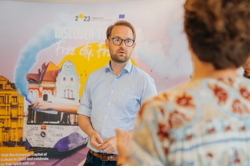 Un bărbat cu ochelari și cămașă albastră deschisă stă în fața unui panou grafic colorat, în timp ce o persoană cu o cămașă modelată este văzută din spate.