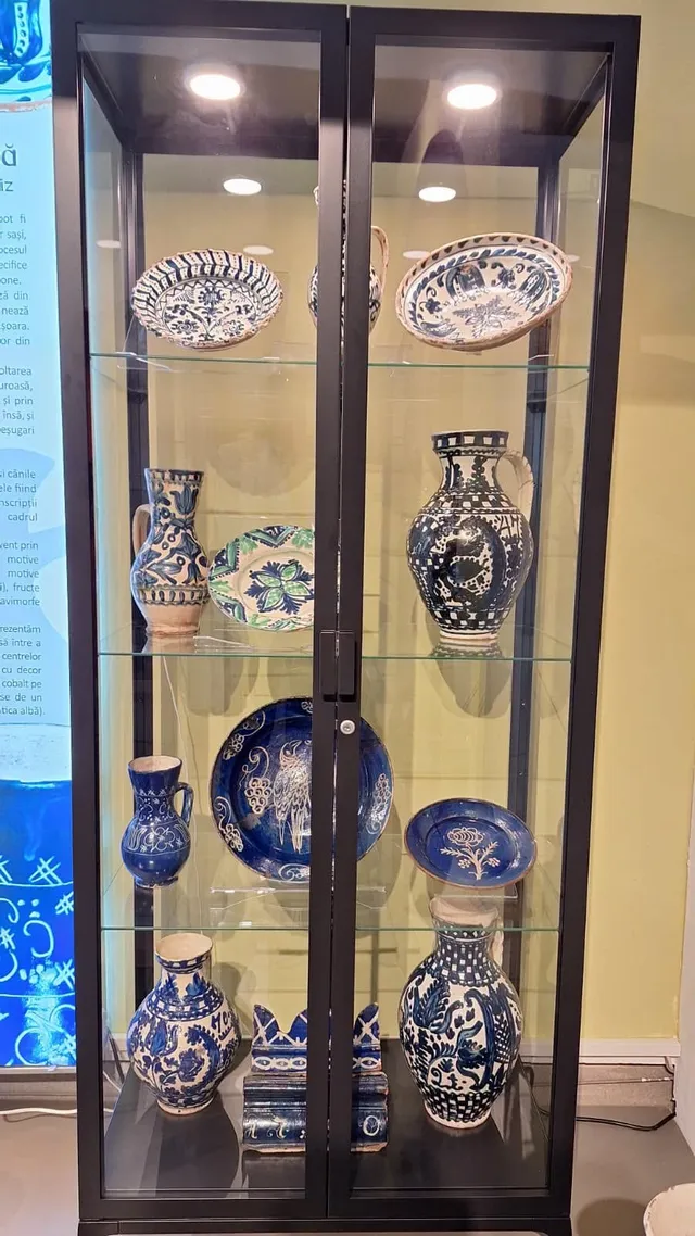 Diverse piese de ceramică albastră și albă, inclusiv vase și farfurii decorate, sunt expuse pe rafturi de sticlă într-o vitrină neagră.