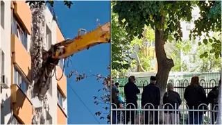 O excavator cu braț galben desface o parte dintr-o clădire, în timp ce un grup de persoane stă în spatele unui gard metalic alb.