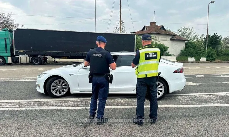 Doi polițiști în uniformă stau lângă un autoturism alb pe o șosea, în fața unui camion mare și al unei clădiri albe.