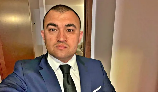 Un bărbat cu părul scurt, purtând un costum albastru, cămașă albă și cravată neagră, privește spre cameră într-un interior cu uși de lemn.