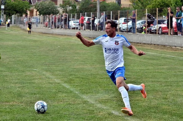Un jucător de fotbal într-un echipament alb și albastru lovește un balon pe un teren de gazon verde.