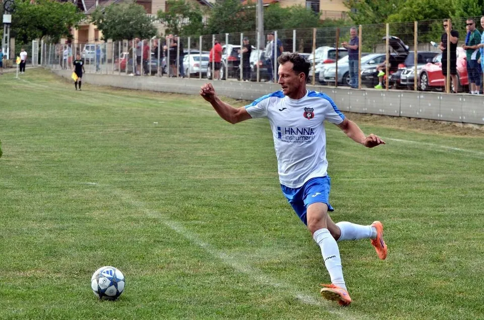 Un jucător de fotbal într-un echipament alb și albastru lovește un balon pe un teren de gazon verde.