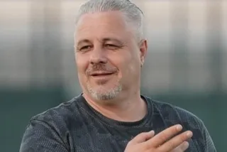 Un bărbat cu părul scurt și gri, purtând un tricou gri închis, zâmbește cu mâna ridicată în prim-plan.