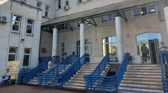 O clădire cu ferestre și trei coloane albe susține un acoperiș, având scări exterioare cu balustrade albastre și câteva persoane în apropiere.