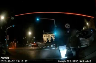 O perspectivă de pe bordul unei mașini surprinde o stradă luminată noaptea, cu un clădire iluminată în fundal și două siluete de lângă vehicul.