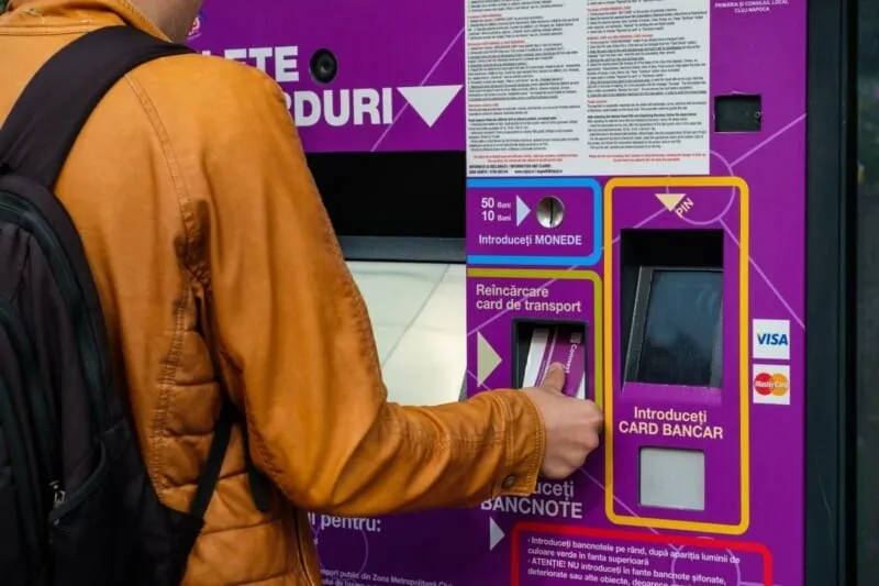 O persoană cu o geantă în spate folosește un automat violet pentru achiziționarea de bilete, introducând un card într-un slot.
