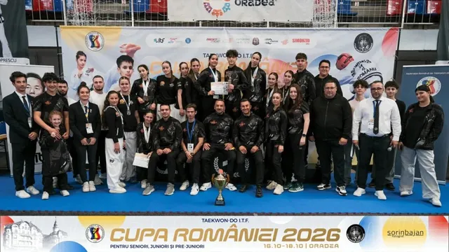 Un grup de persoane îmbrăcate în echipamente sportive negre și haine civile posedă medalii și un trofeu în fața unui panou publicitar colorat.