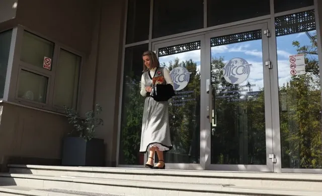 O femeie cu părul blond, purtând o rochie lungă de culoare deschisă și o geantă neagră, pășește pe trepte în fața unei intrări cu uși de sticlă.