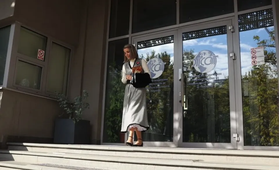 O femeie cu părul blond, purtând o rochie lungă de culoare deschisă și o geantă neagră, pășește pe trepte în fața unei intrări cu uși de sticlă.