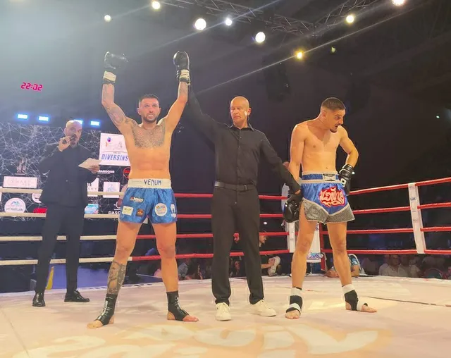 Trei bărbați stau pe un ring de luptă, unul cu mâinile ridicate, în timp ce un arbitrul stă între doi luptători dezbrăcați din partea superioară.