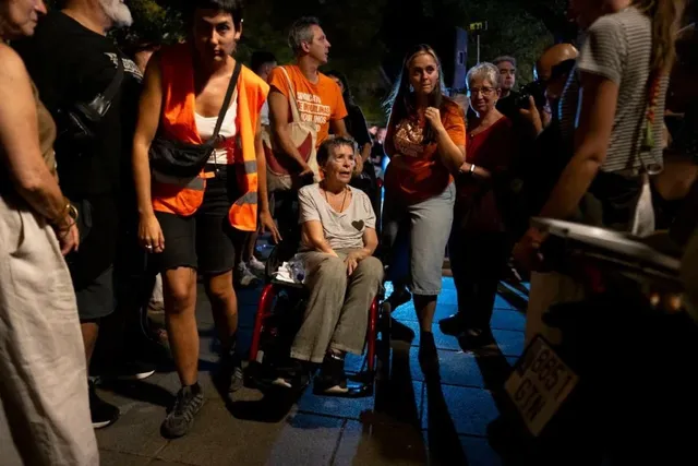 O femeie în vârstă stă în scaun rulant, înconjurată de un grup de oameni pe un drum iluminat noaptea.