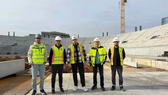 Cinci bărbați cu veste reflectorizante și căști de protecție stau în picioare pe un șantier cu structuri de beton și o macara în fundal.