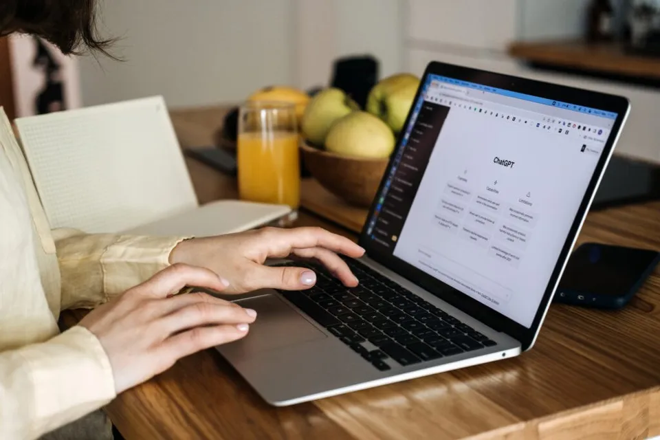 O persoană cu o cămașă deschisă la culoare tastează pe un laptop aflat pe o masă de lemn lângă un notebook și un pahar cu suc.