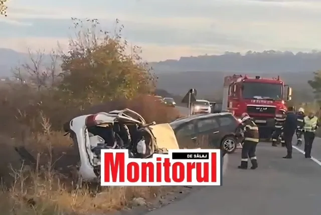 Două autoturisme accidentate stau pe marginea unui drum, lângă un camion roșu și persoane cu uniforme de serviciu.